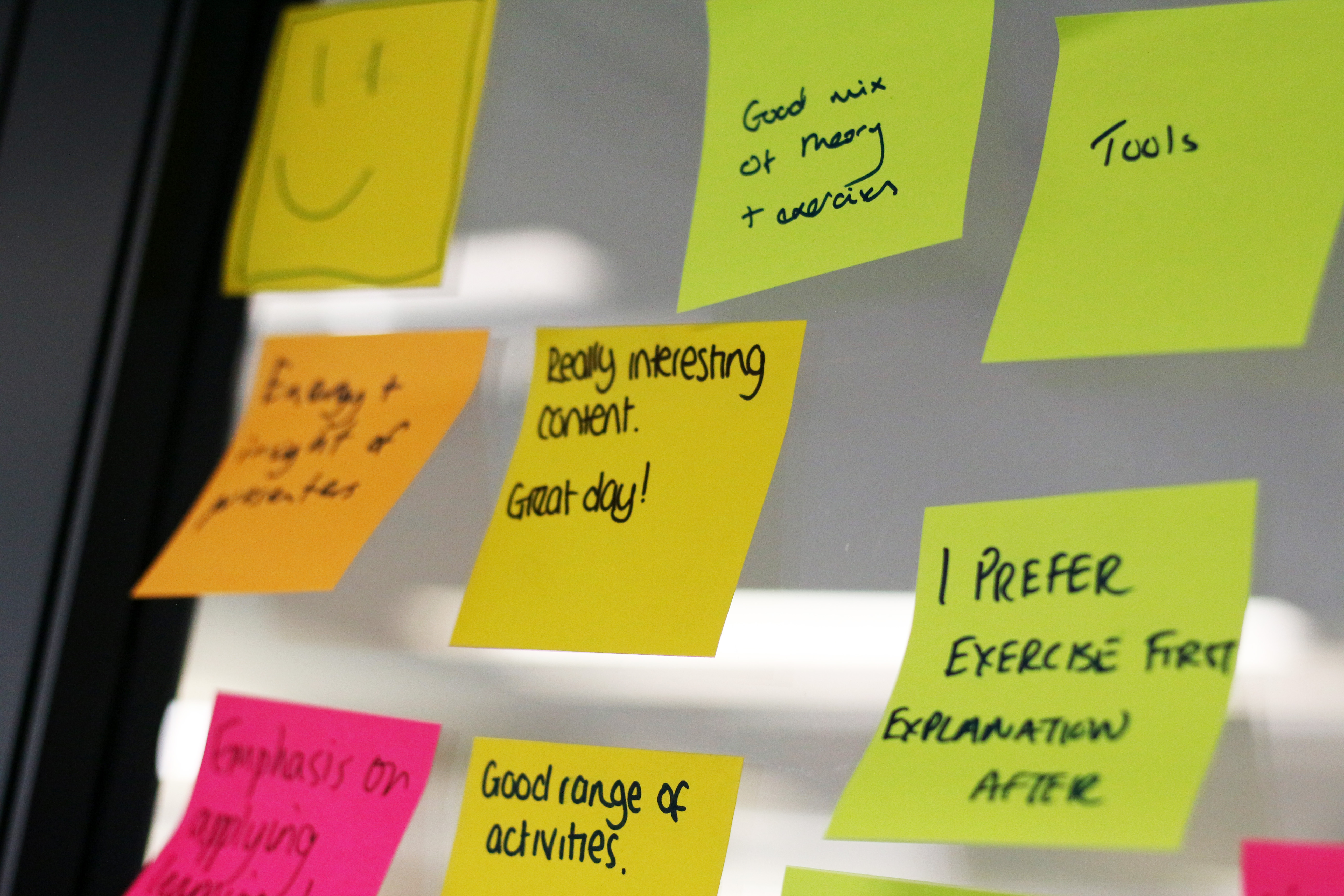 Post-it notes on a window. The ones in focus say "Really interesting content. Great day!", "Good mix of theory + research", "Tools", "Good range of activities." and "I prefer exercise first explanation after""